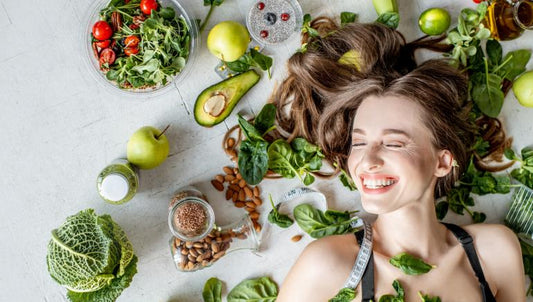 Foto de cima de mulher deita sorrindo com cabelo espalhado e diversos vegetais, frutas, grãos  ao seu redor